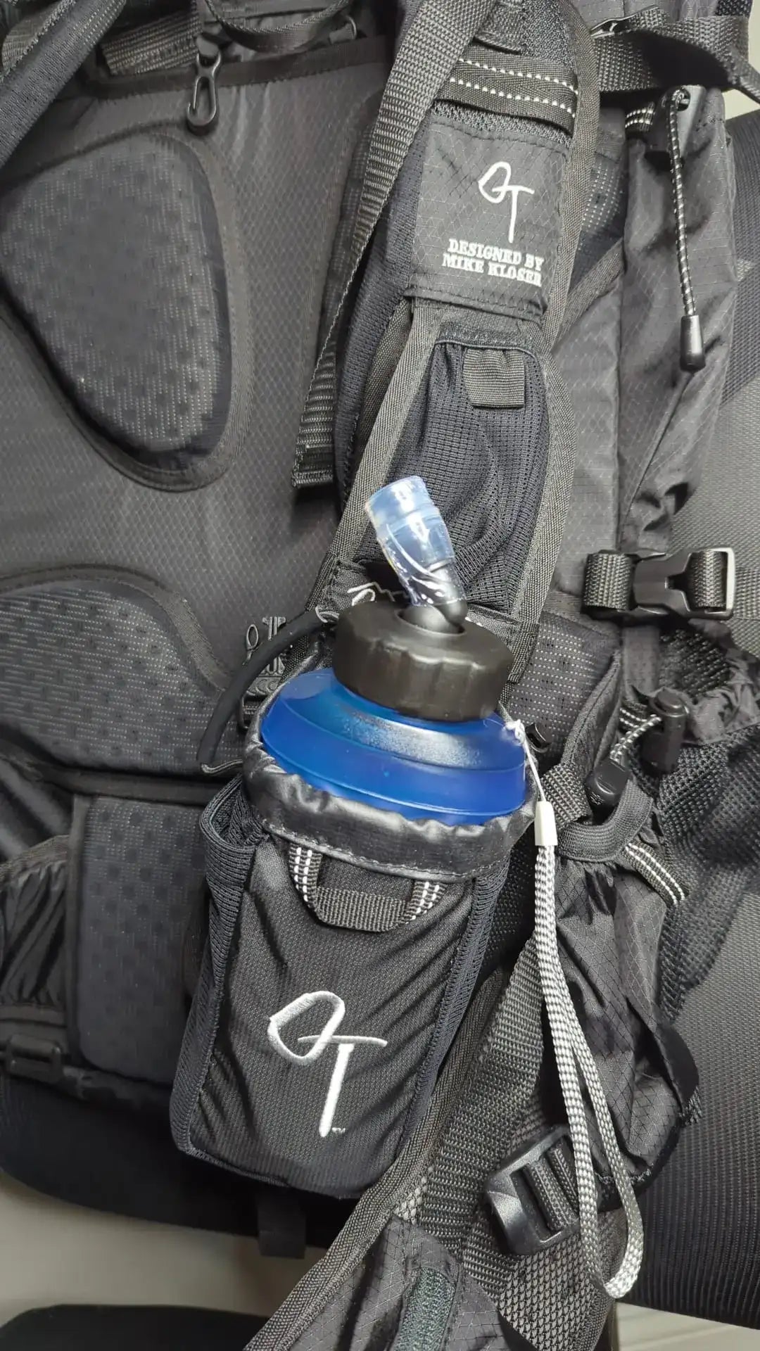 Black backpack with a blue water bottle holder attached, featuring a visible brand logo.